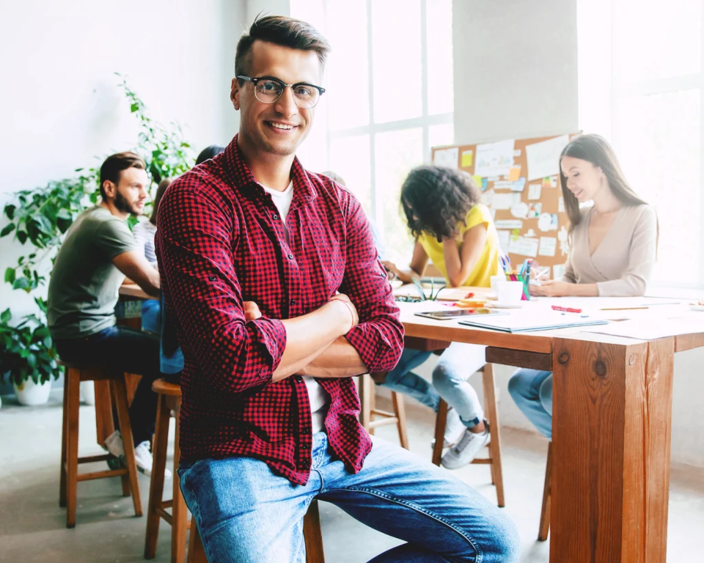 Hombre sentado en frente del aula cruzado de manos sonriendo y grupo de jóvenes estudiantes sentados en mesa de estudio